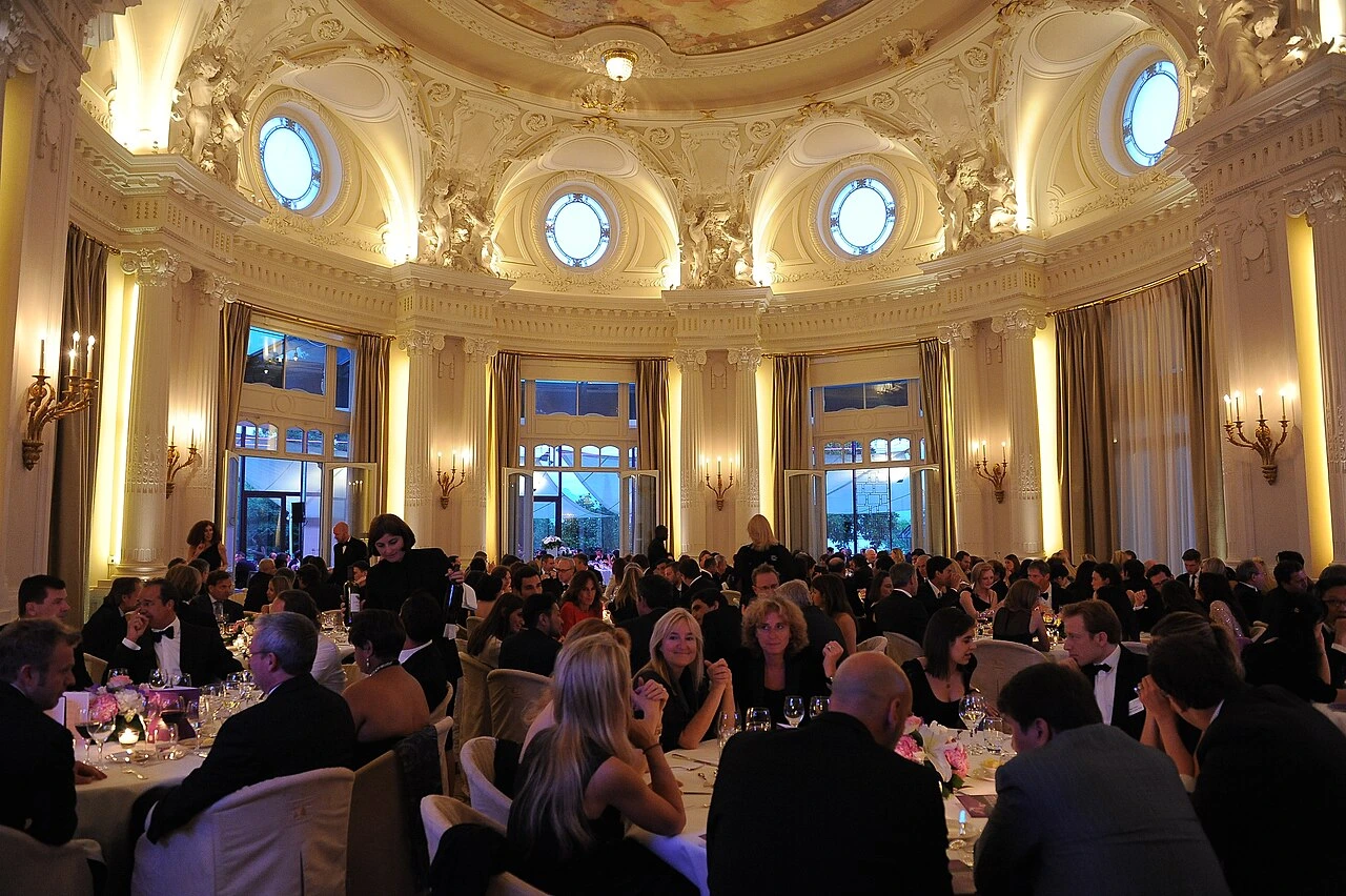 Numerosas personas de etiqueta cenan en mesas redondas en un salón de gala iluminado con luz cálida. Fotografía de referencia.